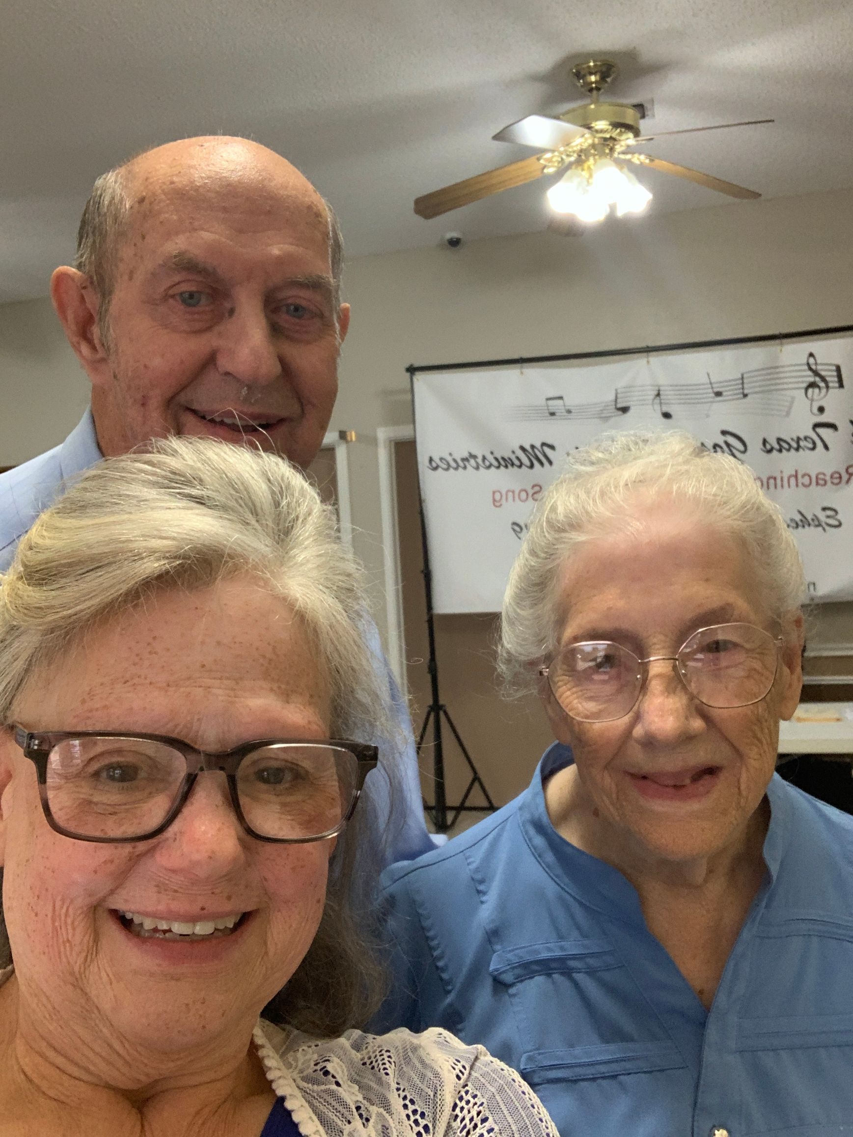 Three people smiling indoors, two women and a man. Blue shirts, eyeglasses, bright lighting.