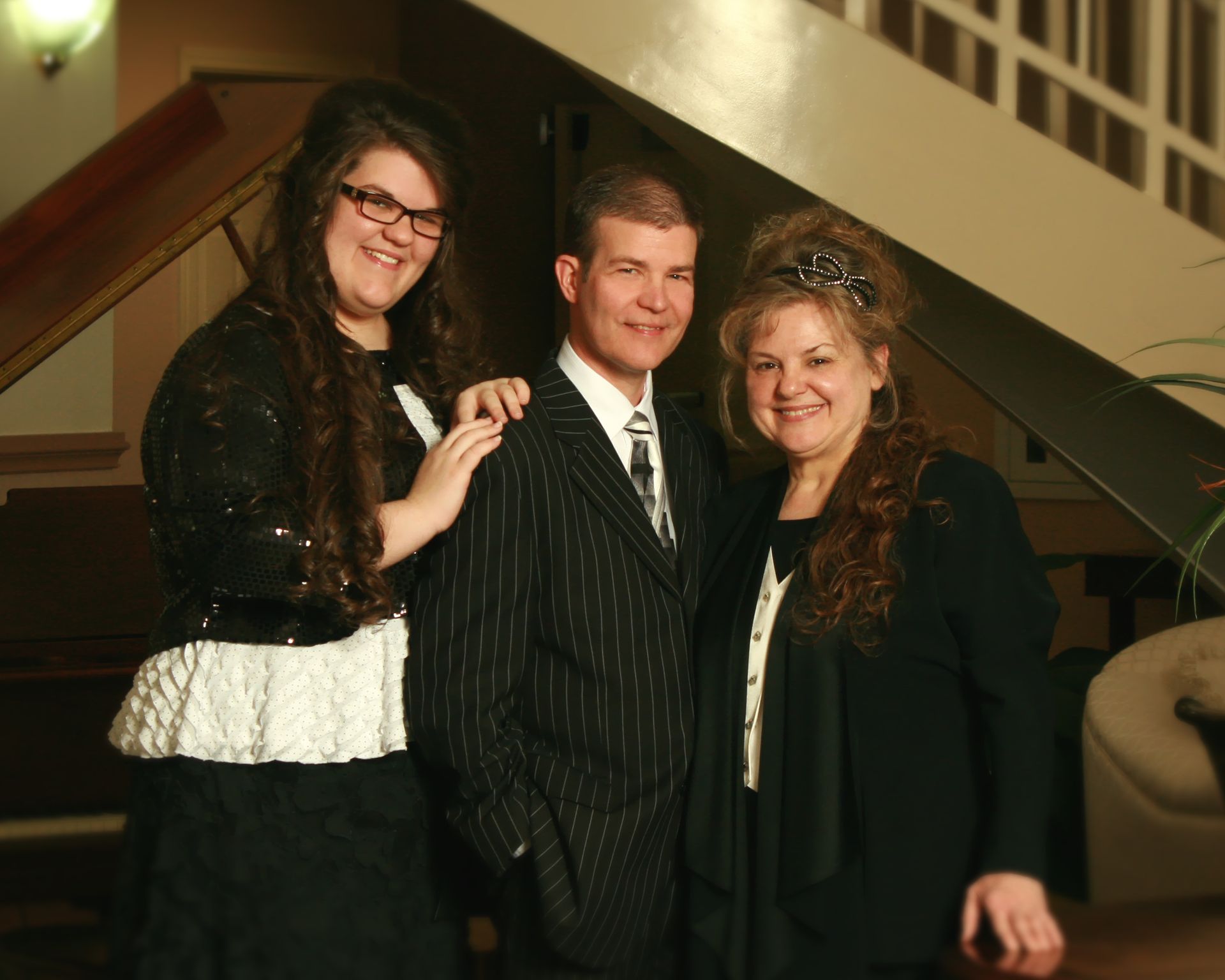 Three people smiling, posed inside near a staircase. The man in pinstripe suit, woman in black, and girl in a sparkly jacket.