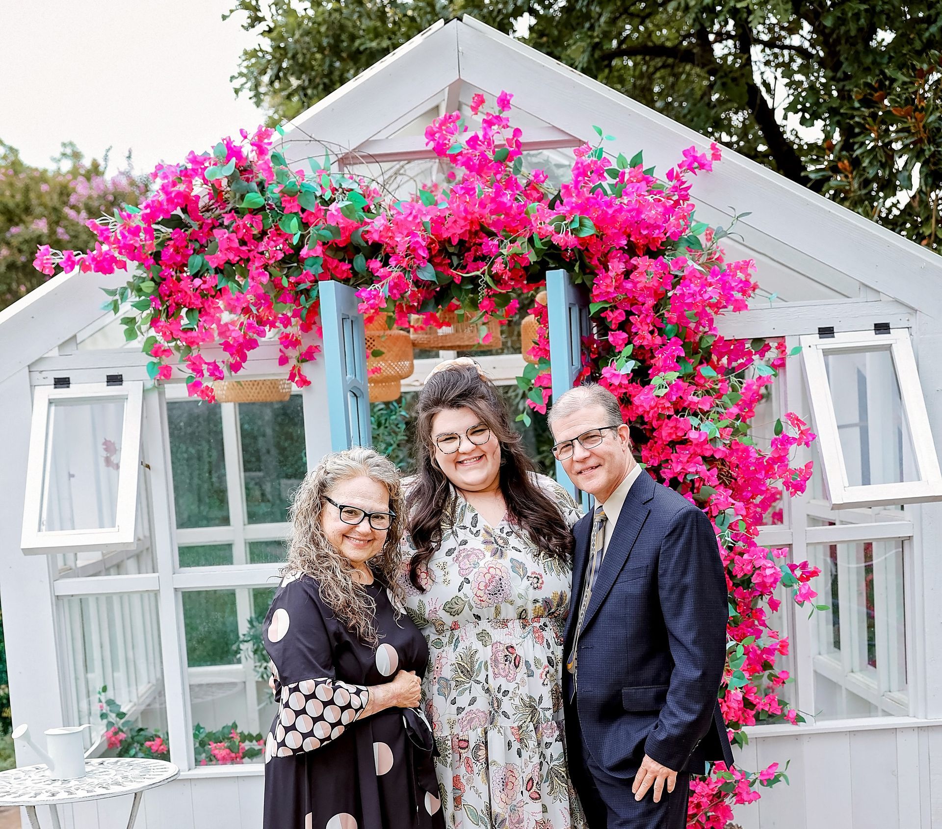 A man and two women are posing for a picture in front of a greenhouse with pink flowers.