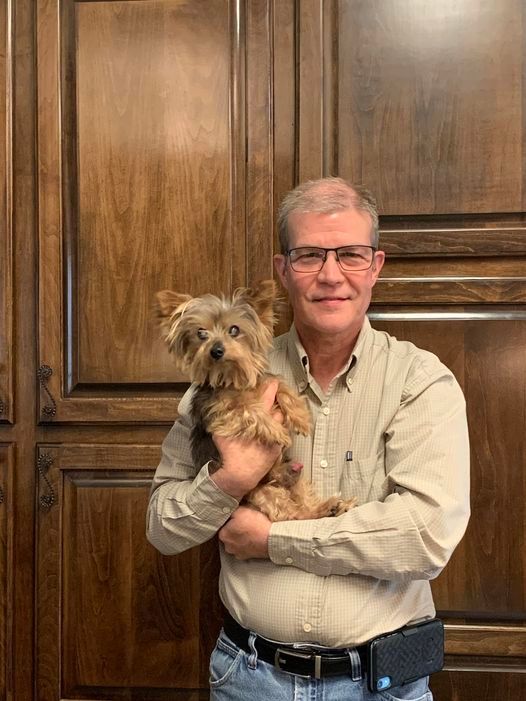 A man is holding a small dog in his arms in front of a wooden wall.