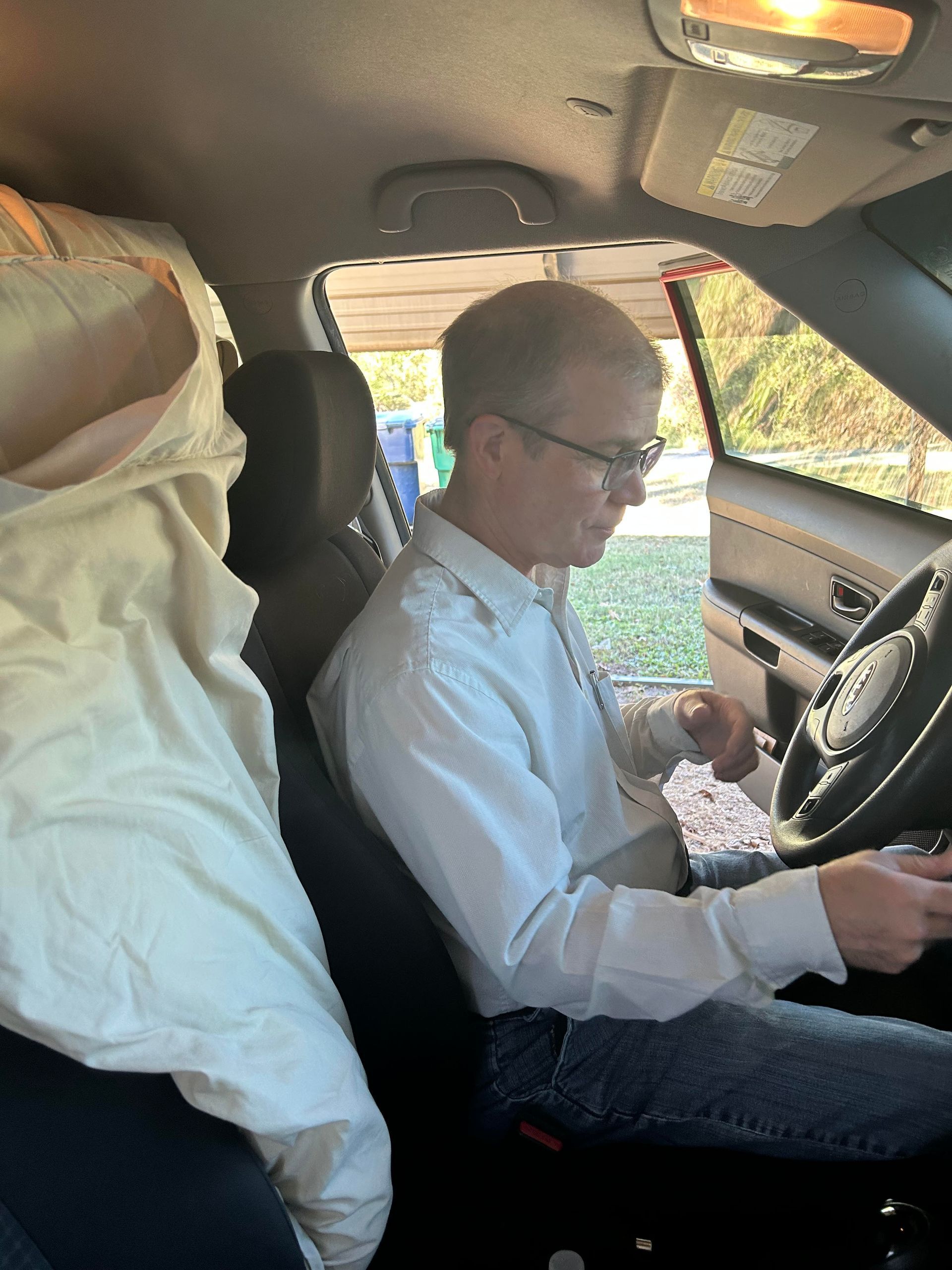 Man sitting in a car, wearing glasses and a light-colored shirt, holding the steering wheel.
