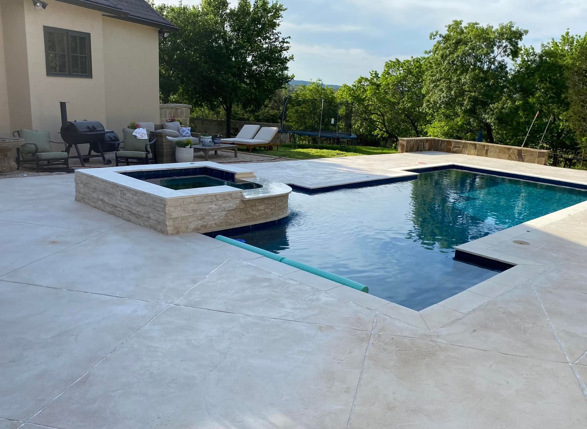 Pool with spa, surrounded by light stone patio. House and trees in background.