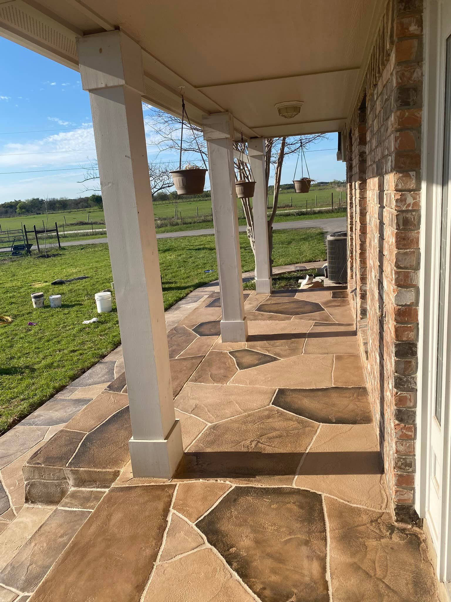 Covered porch with flagstone floor, white columns, and hanging planters; next to brick wall.