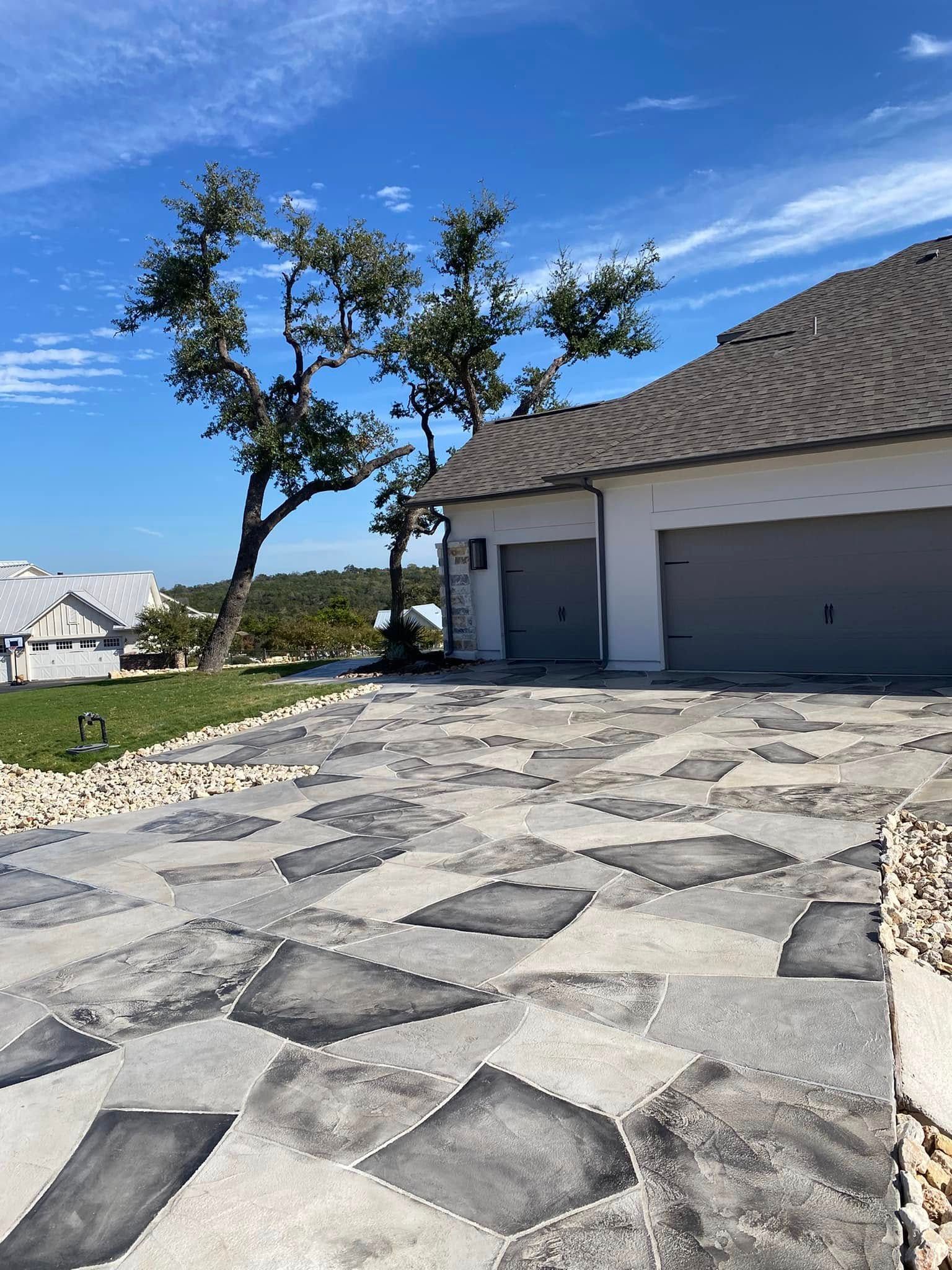 Driveway with patterned stones leading to a white house with grey garage doors; a tree and blue sky in the background.