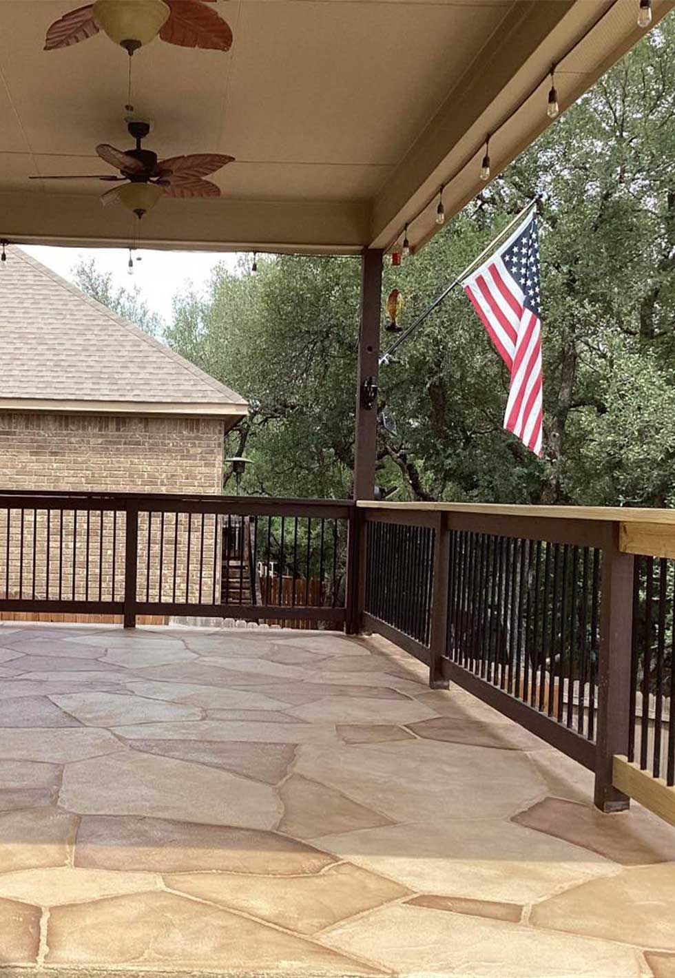 Covered patio with stone floor, dark railing, American flag, ceiling fan, and brick house.