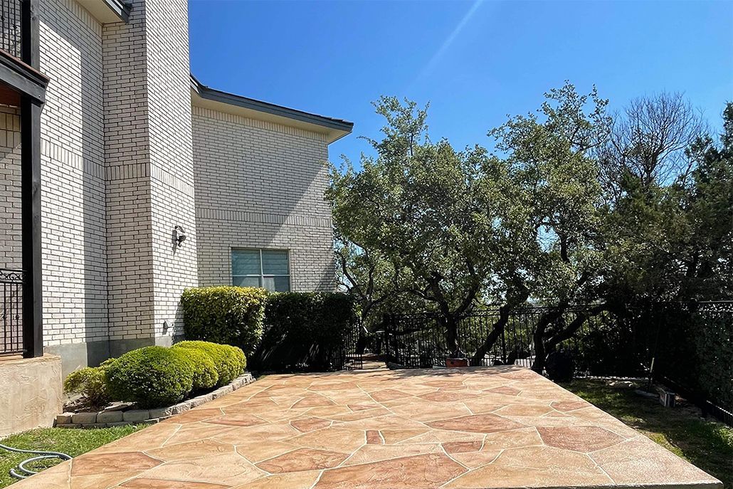 Brick house with stone patio, bushes, and trees under a clear blue sky.