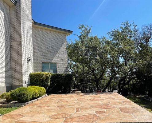 Stone patio beside a brick house, with green bushes and trees under a clear blue sky.