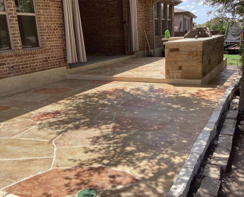 Stone patio with brick house, outdoor kitchen. Beige and red stone, shaded area.