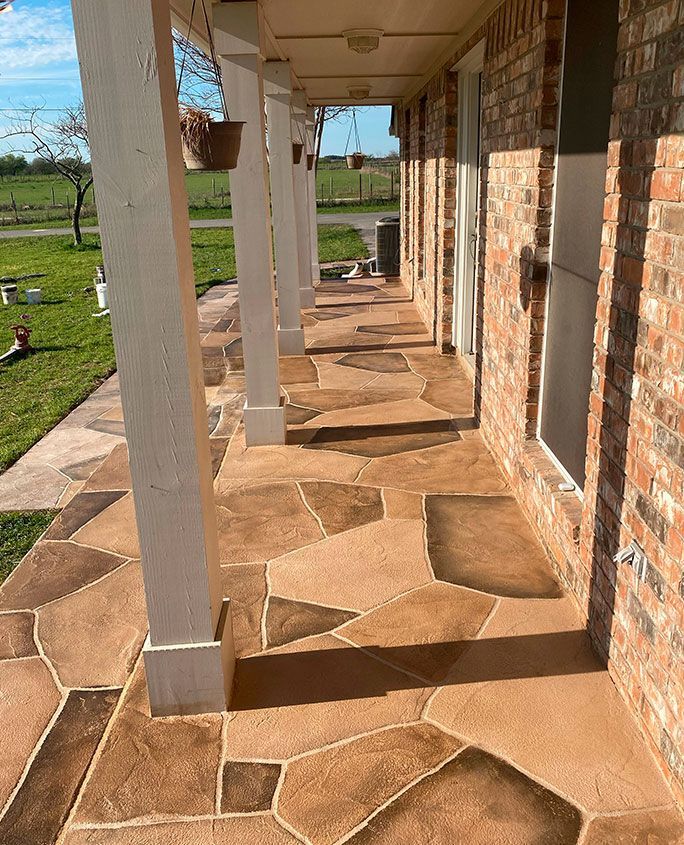 Covered porch with flagstone-like pattern concrete flooring, brick wall, and white columns.