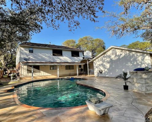 Backyard with pool, patio, and two-story house under a blue sky, trees in the background.