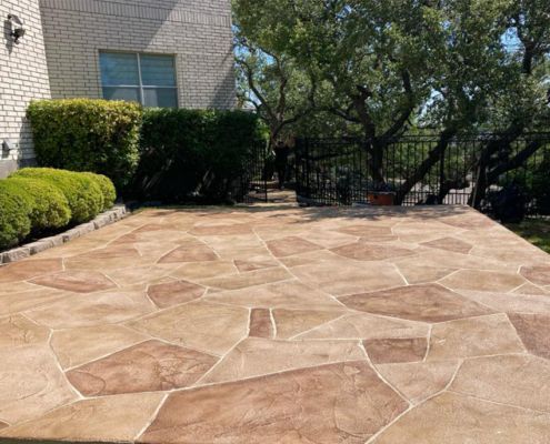 Stone-like patio with brown and tan patterns. Back of a brick home with green bushes and a tree in the background.