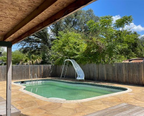 Pool with white slide surrounded by brown stamped concrete, wooden fence, and trees under a blue sky.