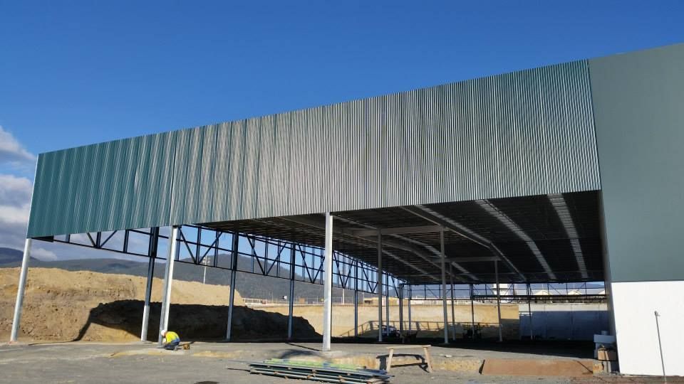 A large metal industrial warehouse under construction, featuring a tall corrugated facade and an open-plan steel frame.