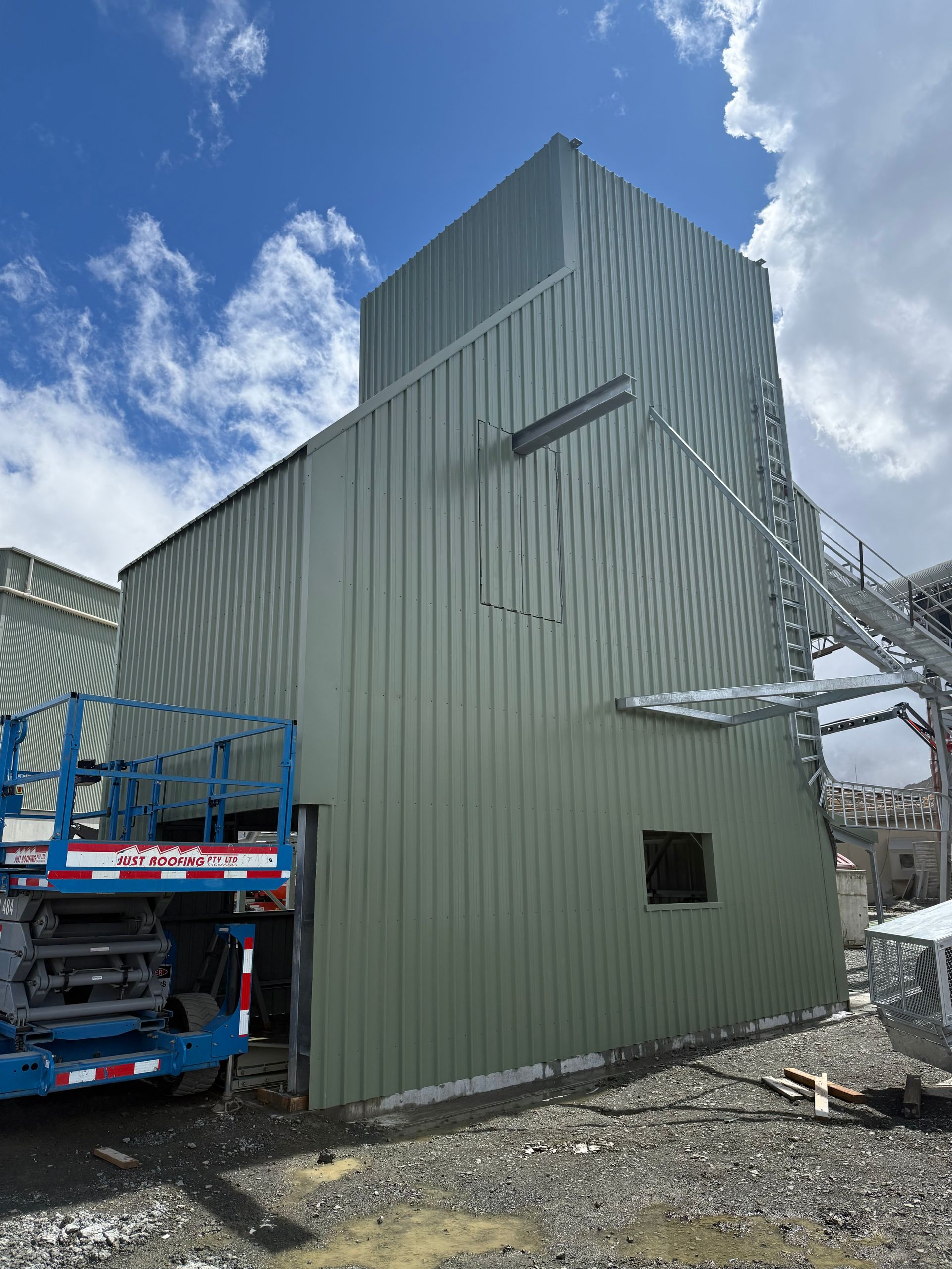 Green metal industrial building beside a blue lift truck under a partly cloudy sky