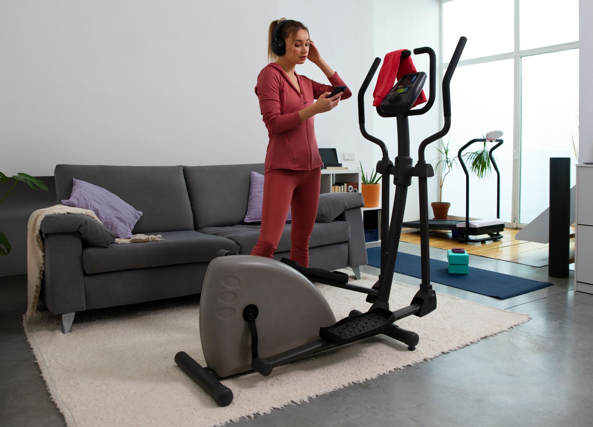 Woman in workout clothes using a phone near an elliptical machine in a living room, with a couch and other fitness equipment.