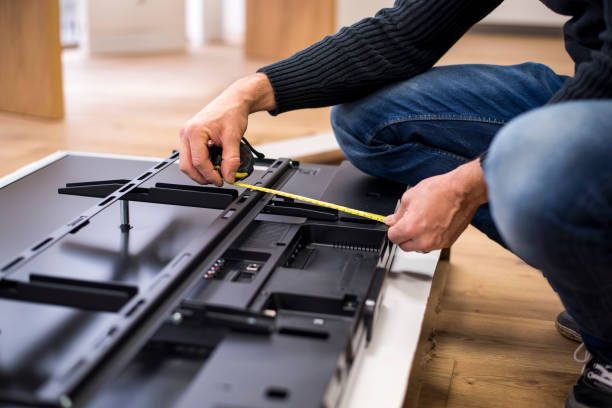 Person assembling a flat-screen TV on a wooden floor with tools and measuring tape