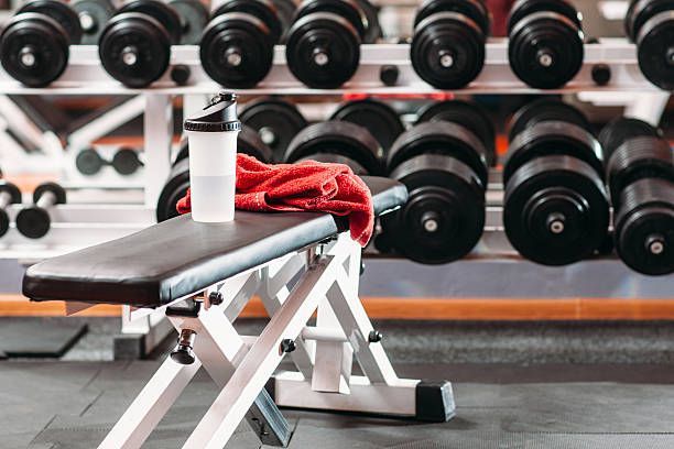 A shaker bottle and a red towel sit on a weight bench in a gym with racks of dumbbells in the background.