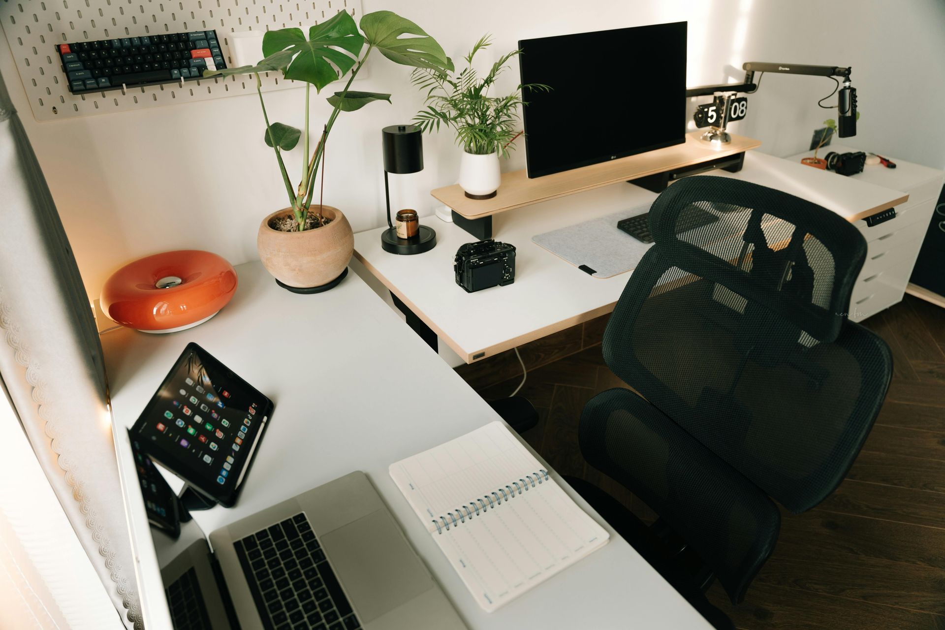 Minimal white desk setup with laptop, monitor, plants, camera, and black mesh chair