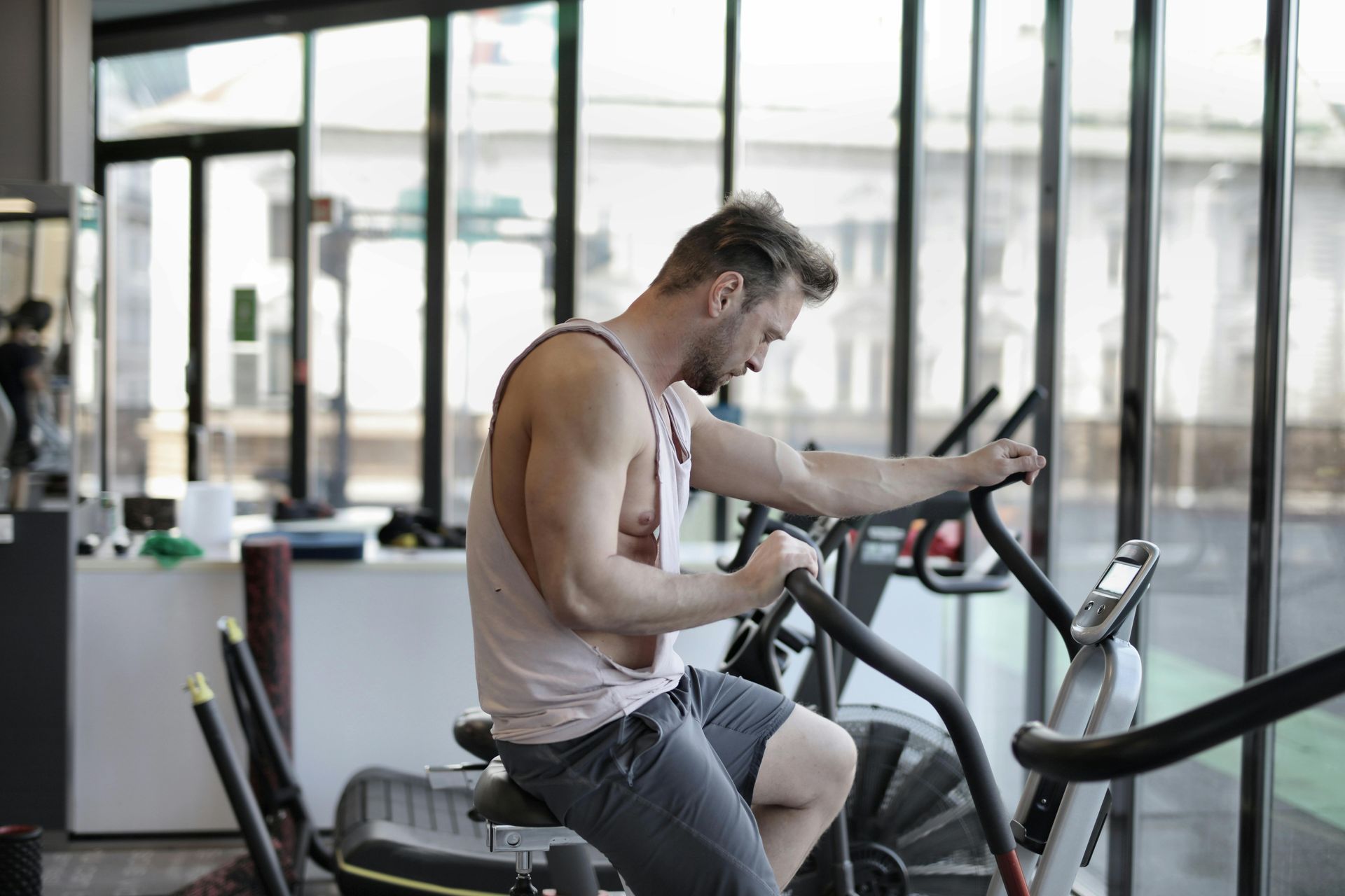 A person in a light tank top and dark shorts exercising on a stationary bike in a gym with large floor-to-ceiling windows.