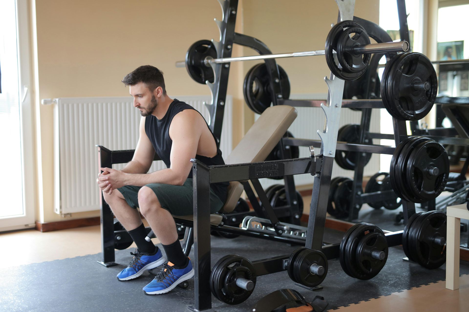 A person in a tank top and shorts sits on a gym bench, looking down with clasped hands next to a weight rack.