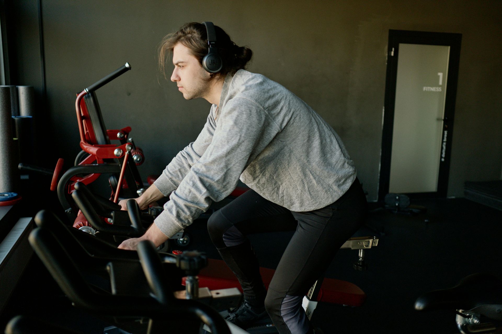 Person exercising on a stationary bike in a gym, wearing headphones and a gray sweater.