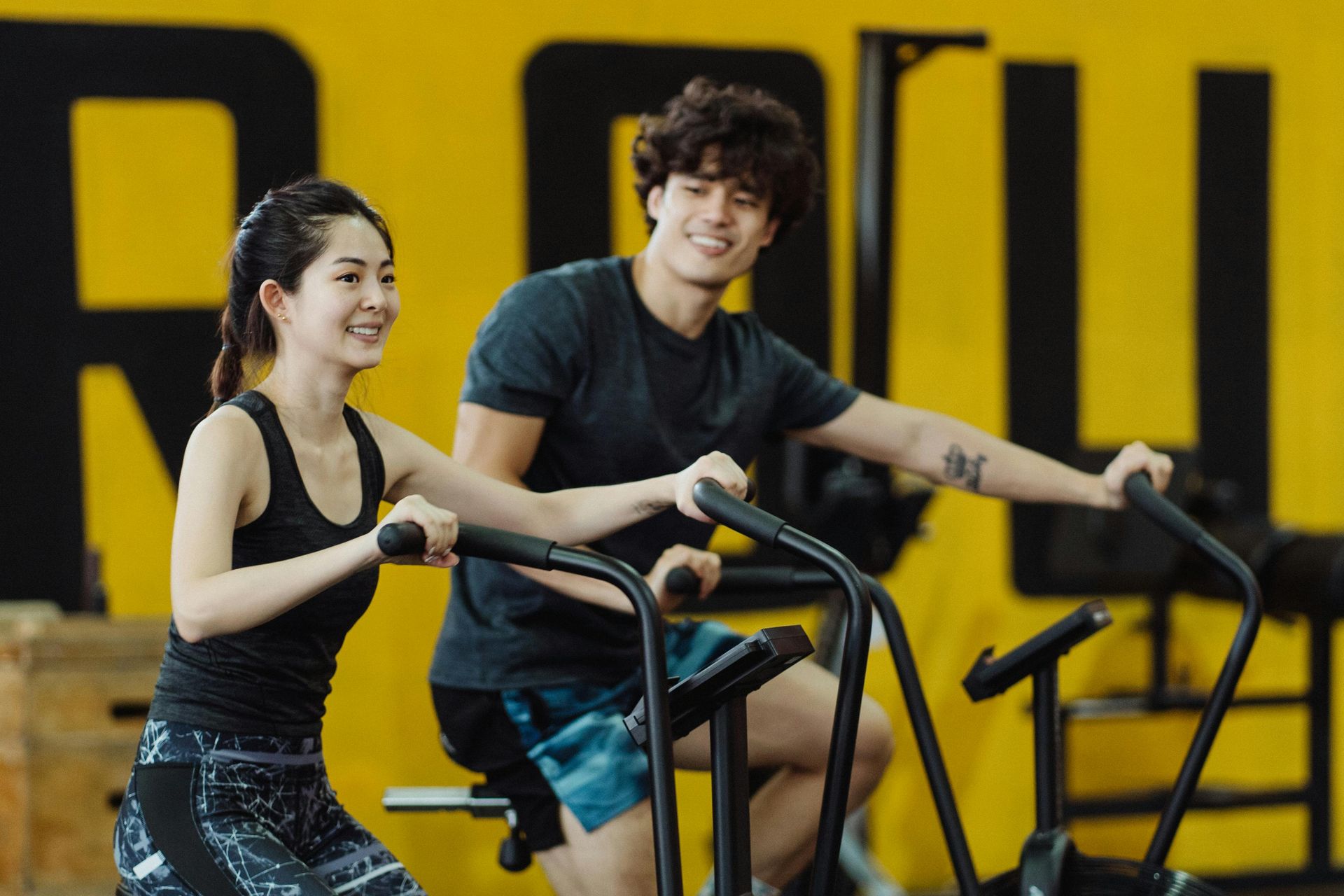Two people exercising together on stationary bikes at a gym with a yellow wall in the background.