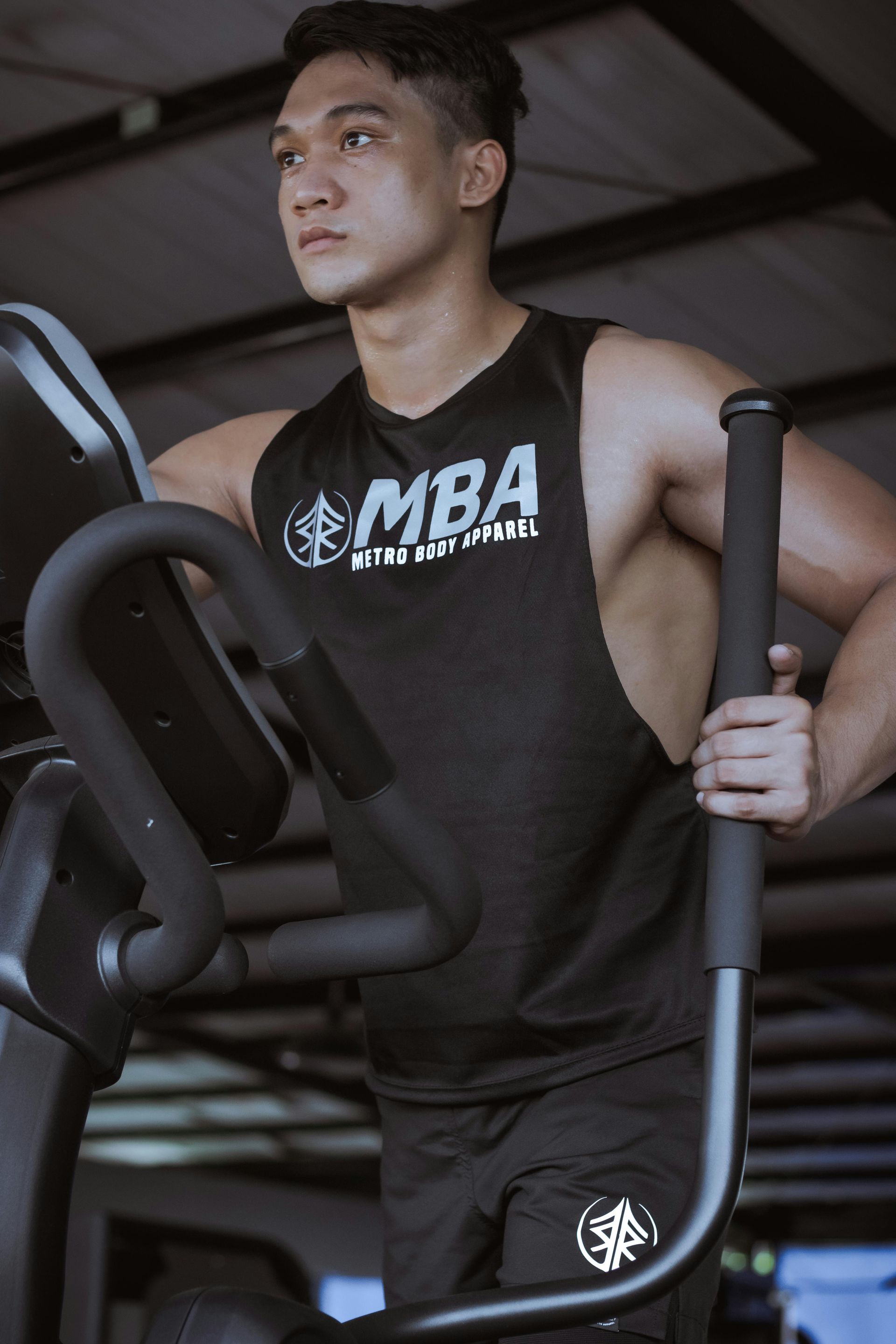 A focused person wears a black muscle tank top while exercising on an elliptical machine at a gym.