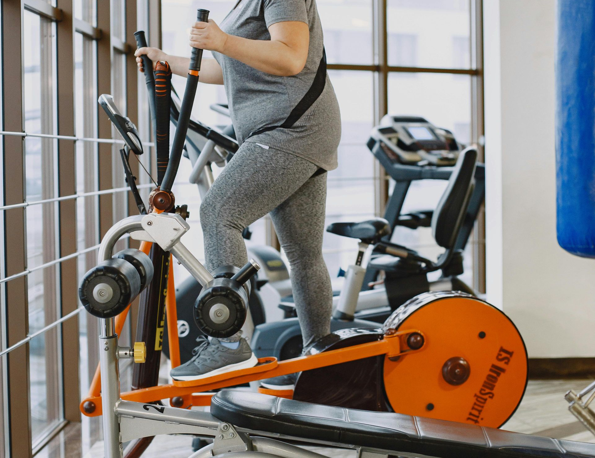 A person in gray activewear exercises on an orange elliptical machine inside a bright, sunlit fitness center.