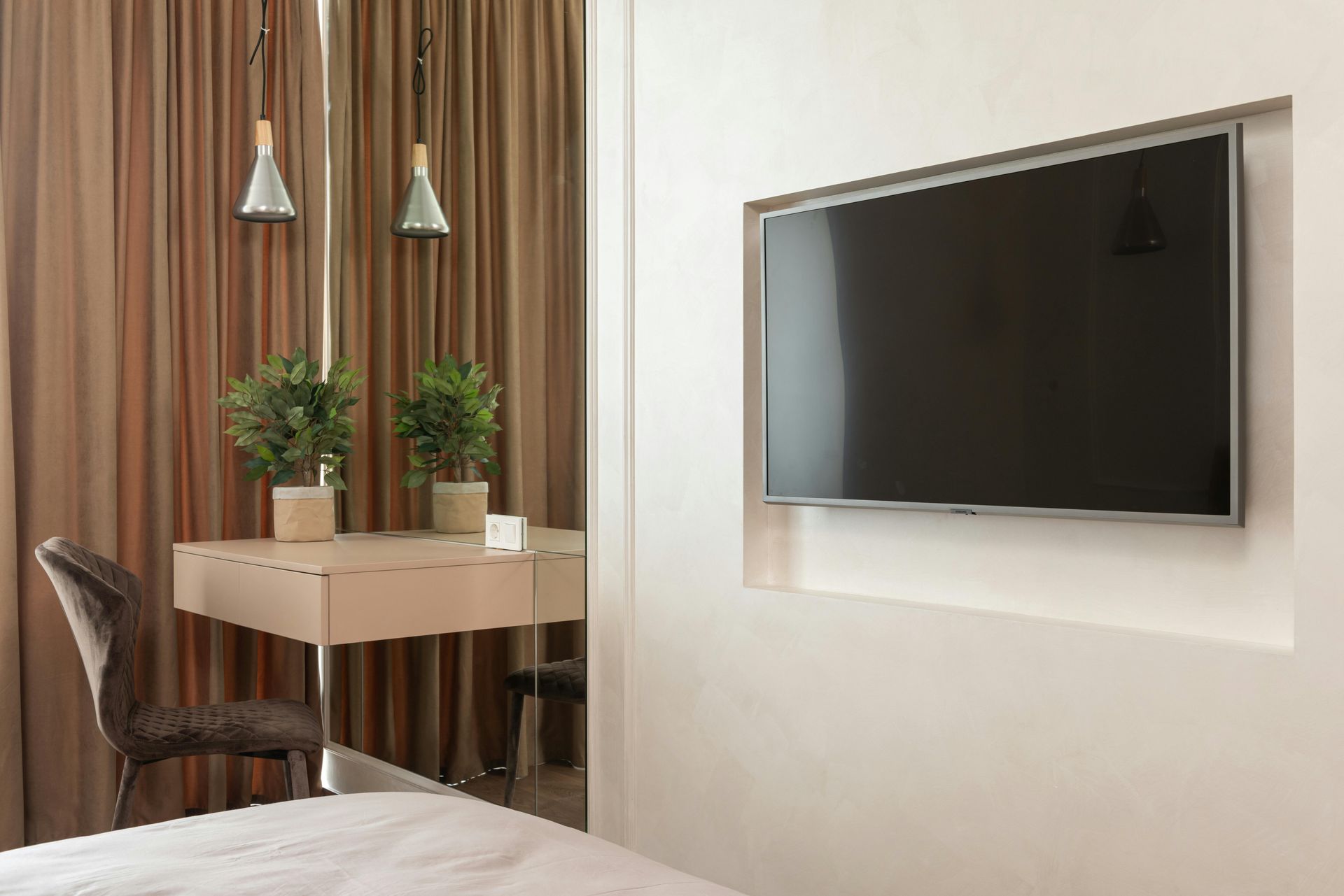 Modern hotel room with a wall-mounted TV, desk, chair, lamp, and potted plants by the window.