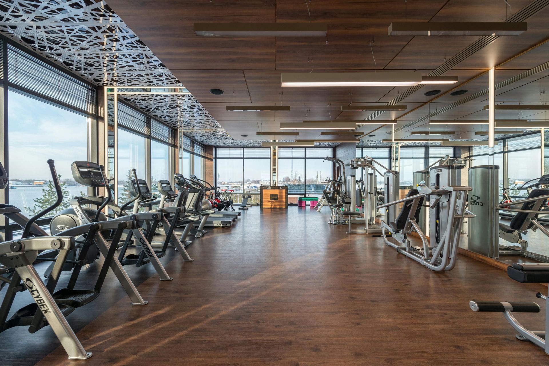 Modern fitness center with rows of gym equipment, wood flooring, decorative ceiling panels, and floor-to-ceiling windows.