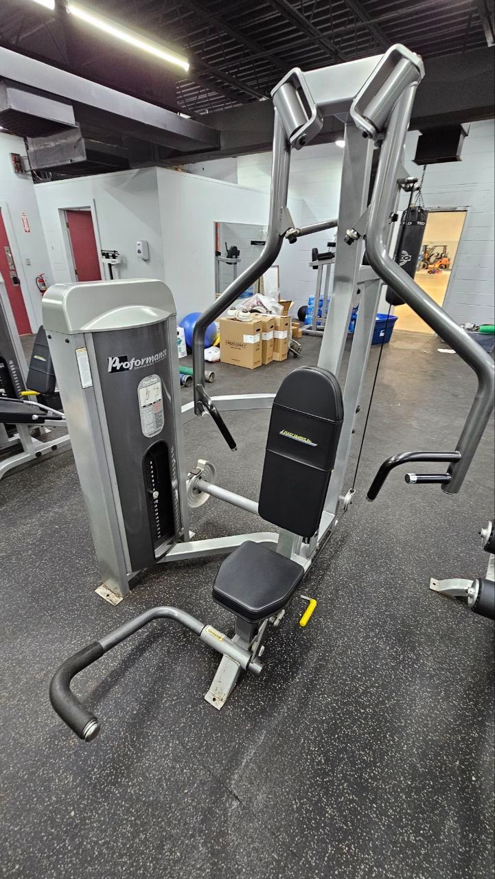 A silver and black chest press machine stands in the center of a gym with rubber flooring.