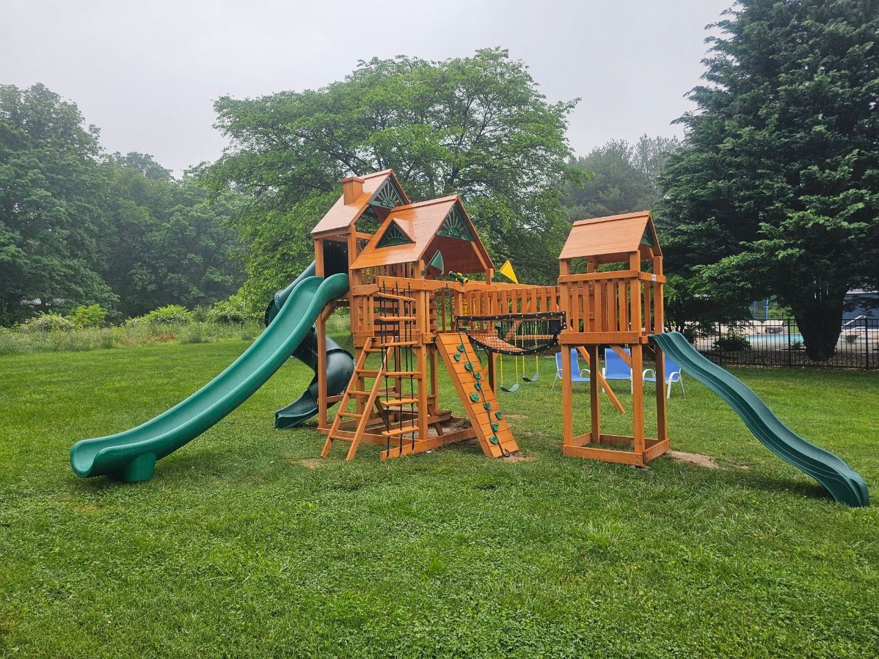 Wooden playset with two green slides, surrounded by grass and trees.