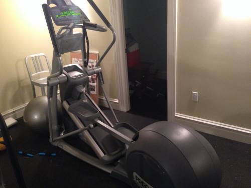 An elliptical Precor machine is sitting in a room next to a chair and a ball.