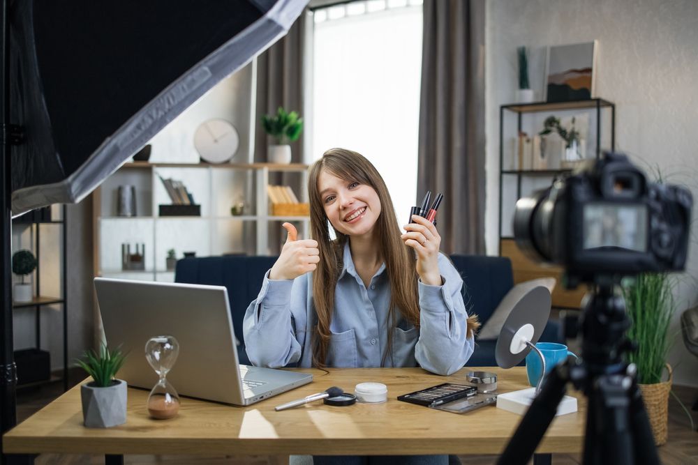 A woman in a blue shirt, holding several different makeup products in one hand, poses with a 'thumbs up'. She is sitting at a desk with an open laptop and a digital camera recording her. What is UGC? The Power of User-Generated Content