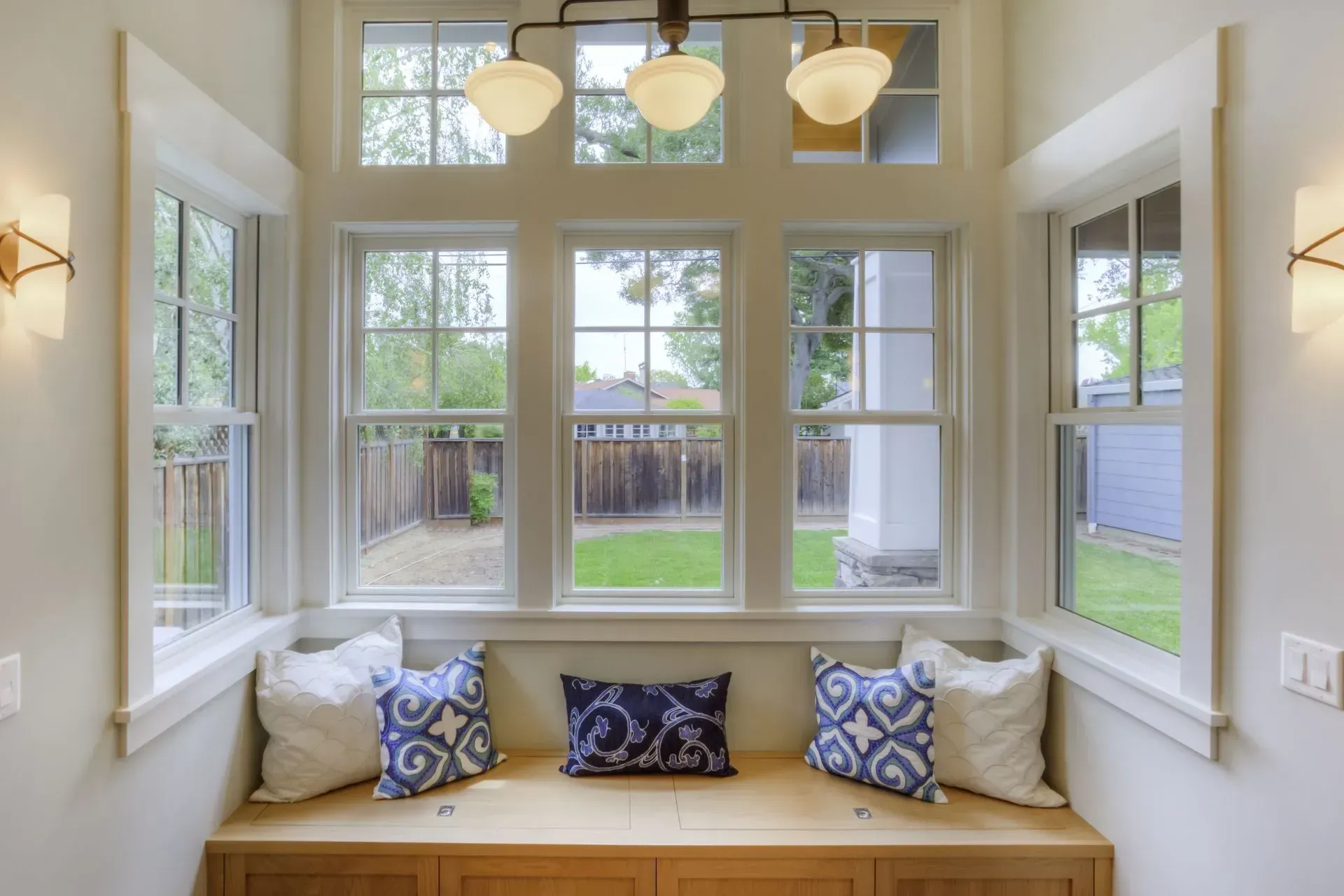 Bright window seat with white trim, blue patterned pillows, and a view of a backyard through large windows