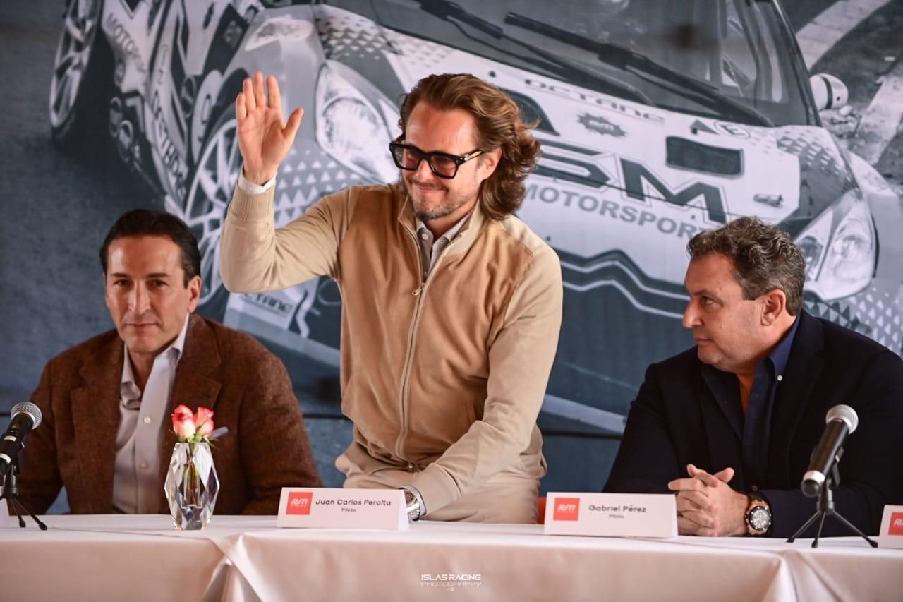 Tres hombres están sentados en una mesa con micrófonos frente a un automóvil.