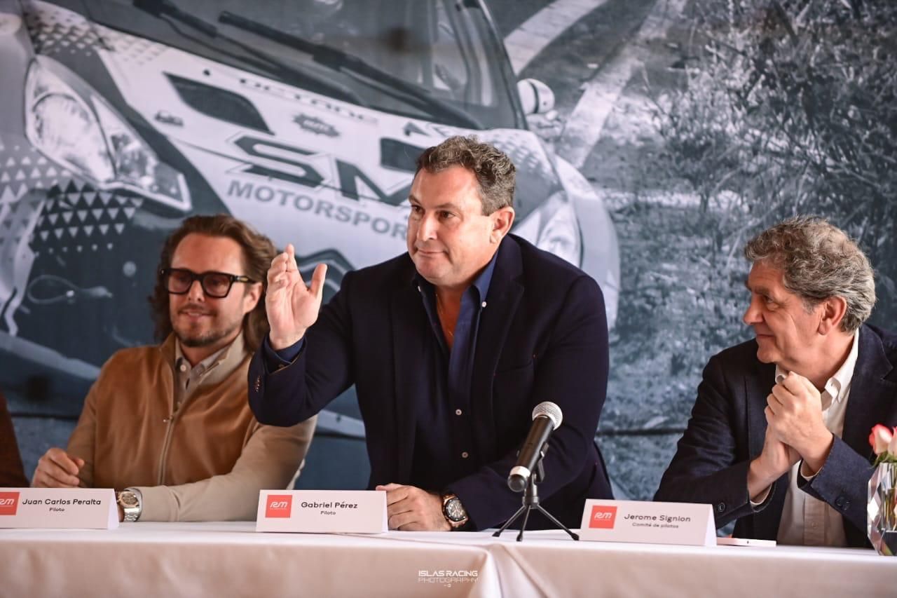 Tres hombres están sentados en una mesa con micrófonos frente a un automóvil.