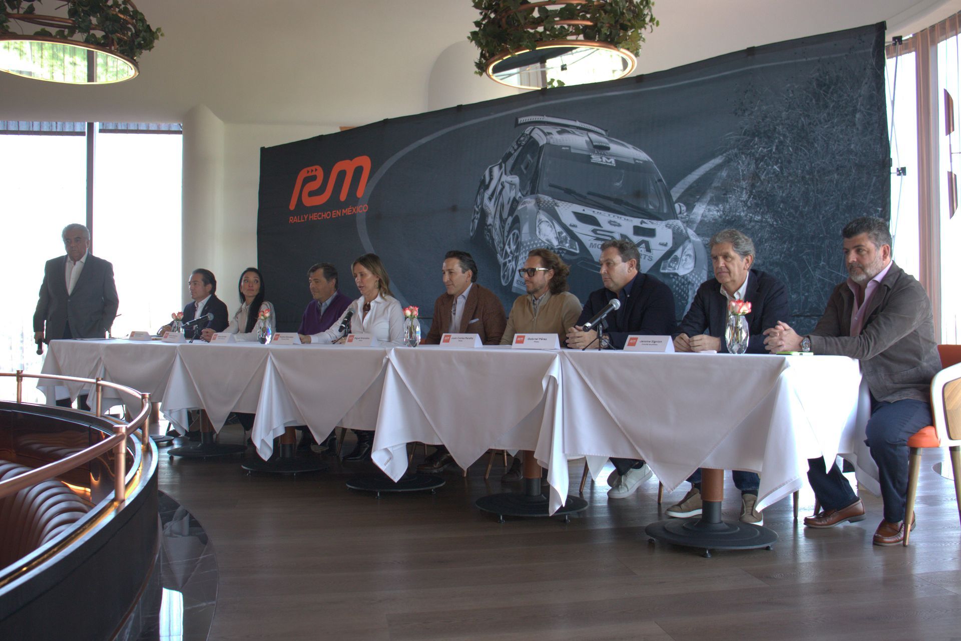 Un grupo de personas están sentadas en mesas frente a una pared que dice rm