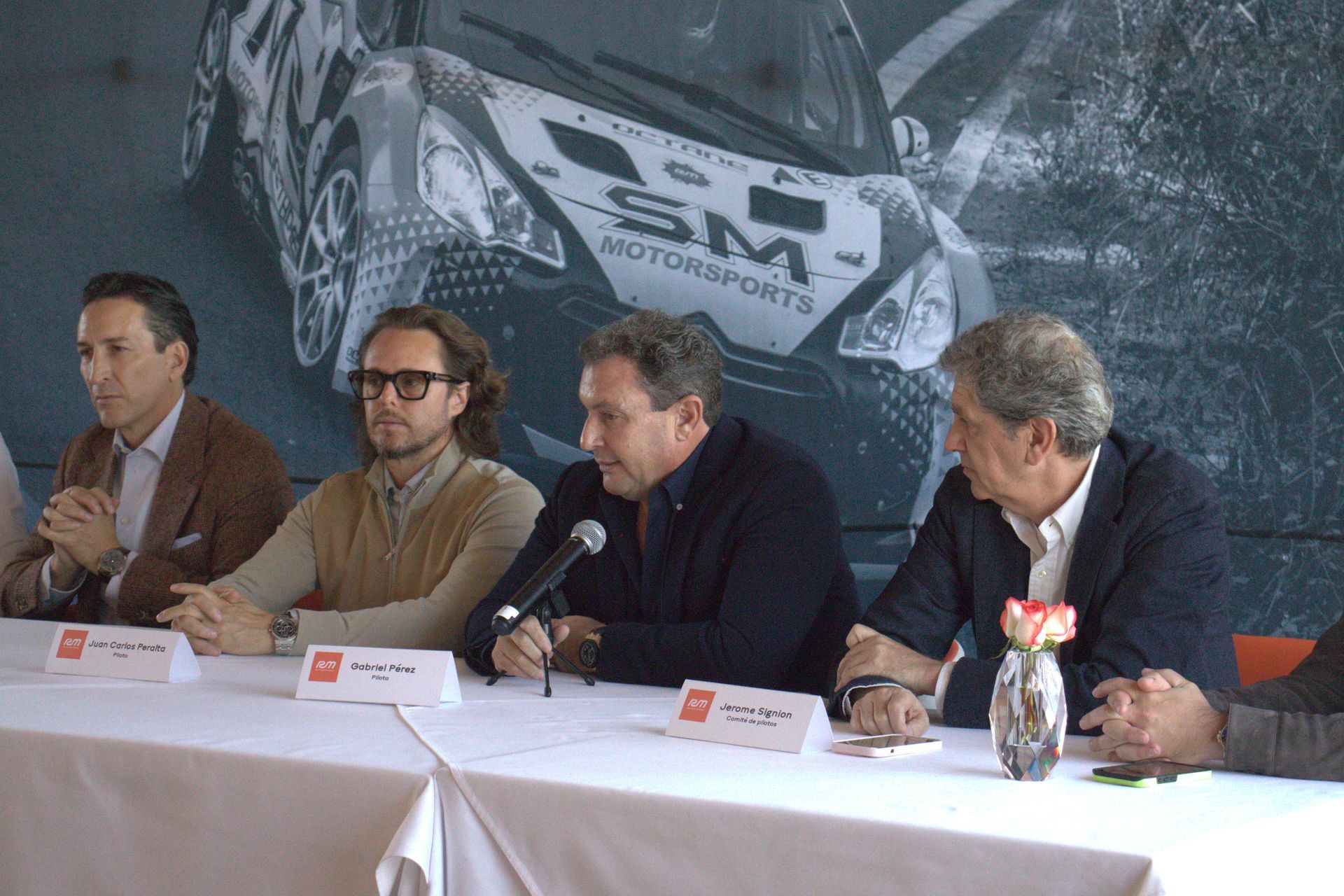 Un grupo de hombres están sentados en una mesa con un automóvil al fondo que dice SM Motorsports.