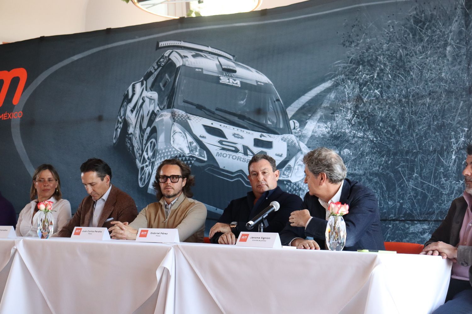 Un grupo de personas sentadas en una mesa con una foto de un automóvil detrás de ellas.