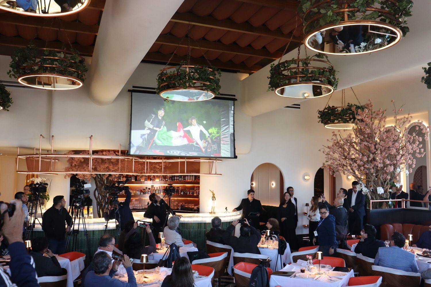 Un grupo de personas están sentadas en mesas frente a una pantalla grande en un restaurante.
