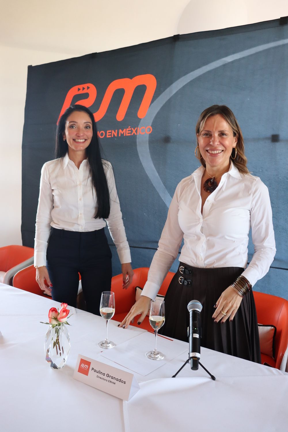 Dos mujeres están sentadas en una mesa con copas de vino y un micrófono.