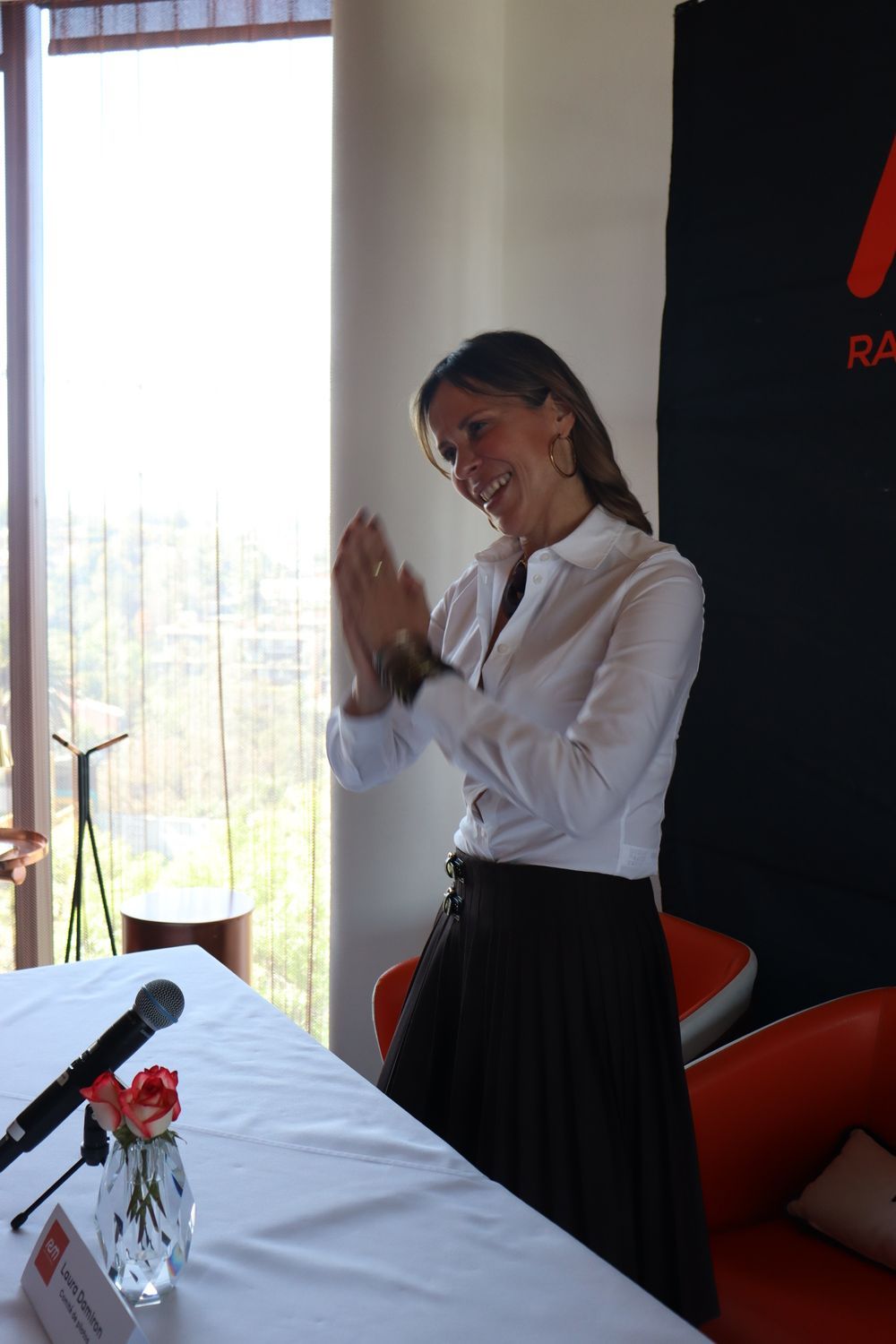 Una mujer con una camisa blanca y una falda negra está sentada en una mesa con un micrófono.