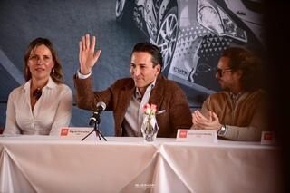 Un hombre agita la mano mientras está sentado en una mesa con otras dos personas.