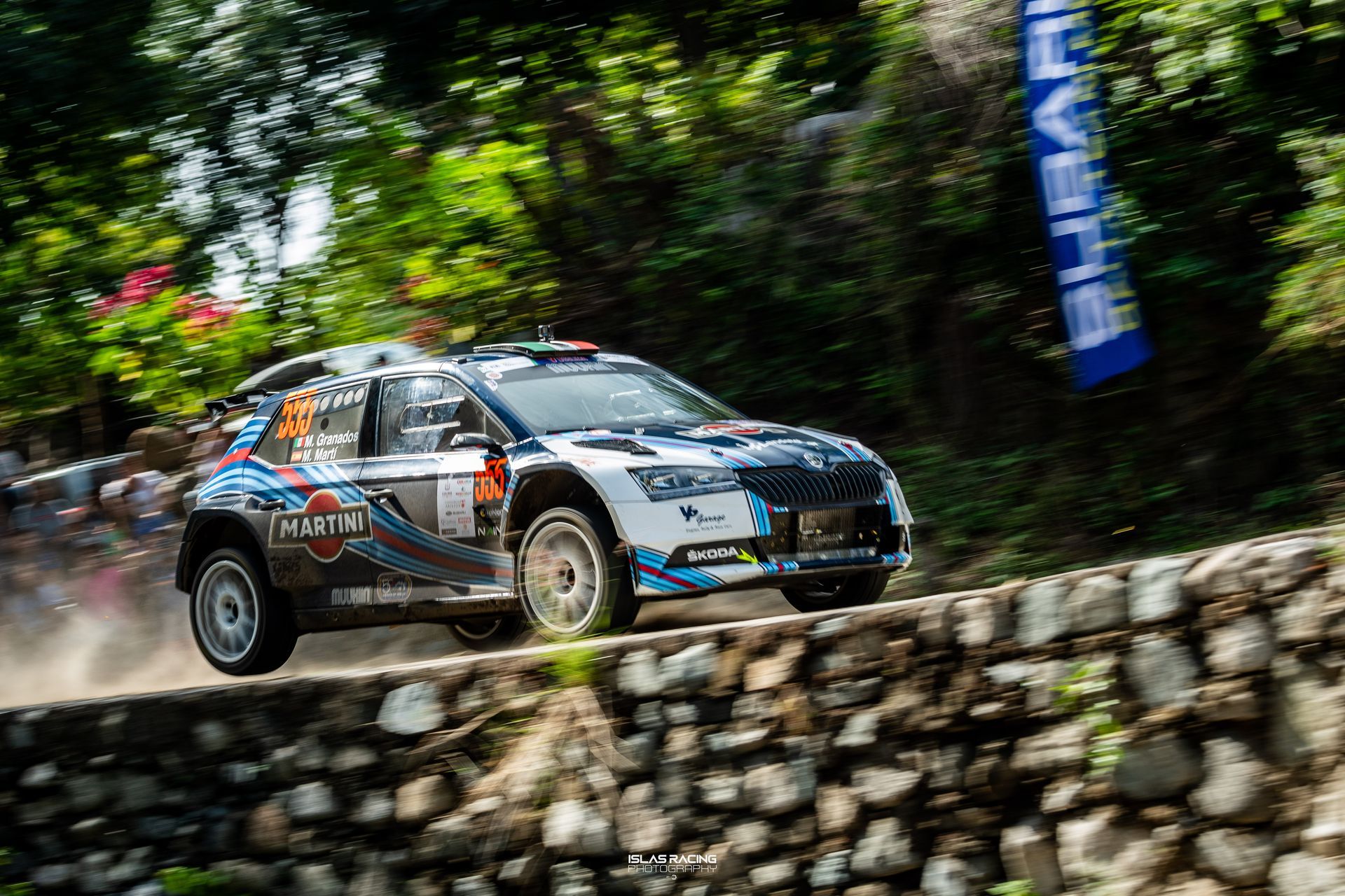 Un coche de rally circula por un camino de tierra junto a un muro de piedra.