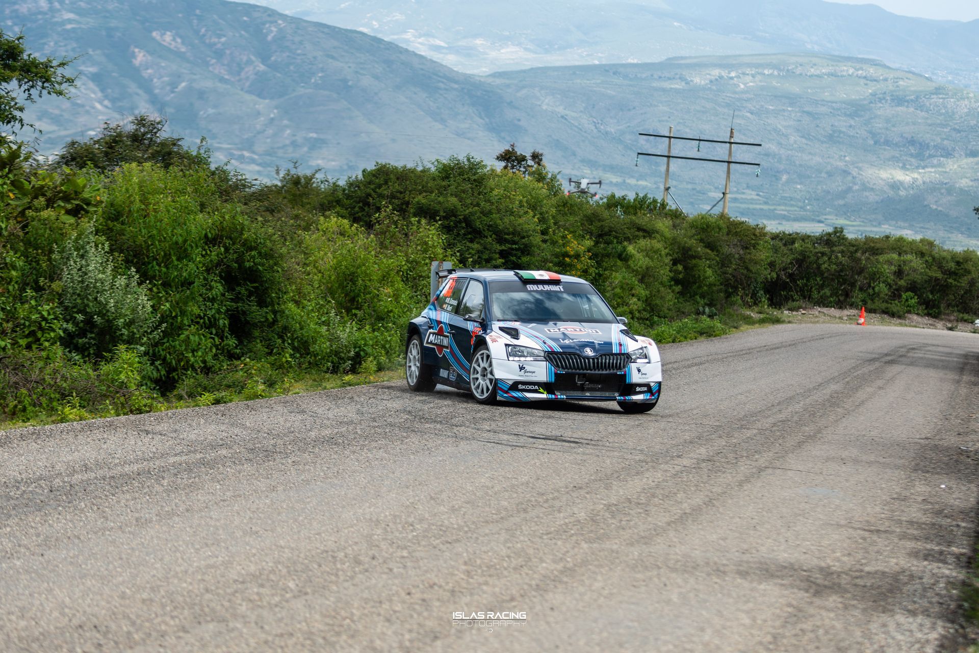 Un coche de rally circula por un camino de tierra con montañas al fondo.