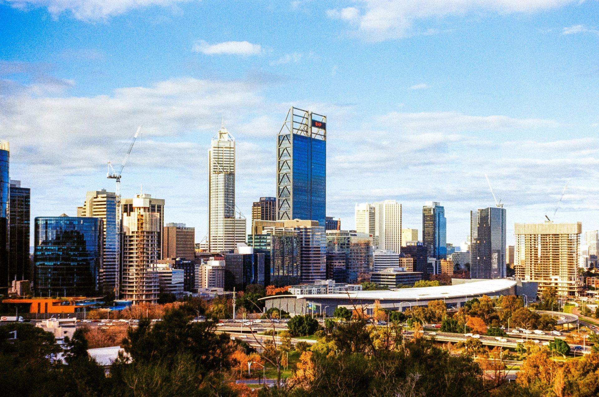City skyline with tall modern buildings under a blue sky, viewed above trees and a highway.