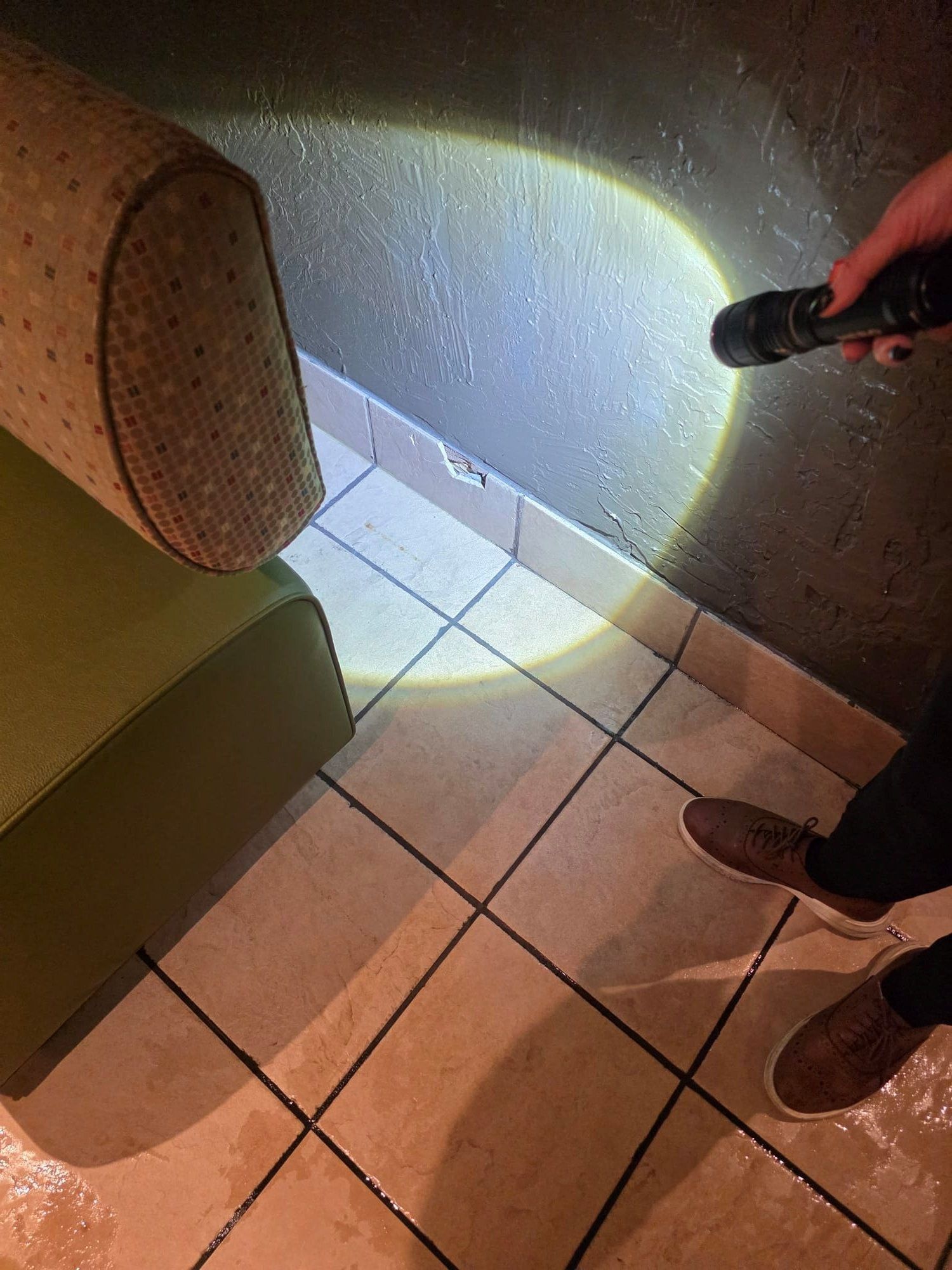 Person shines flashlight on wall near a green couch and tiled floor, possibly inspecting for water damage.