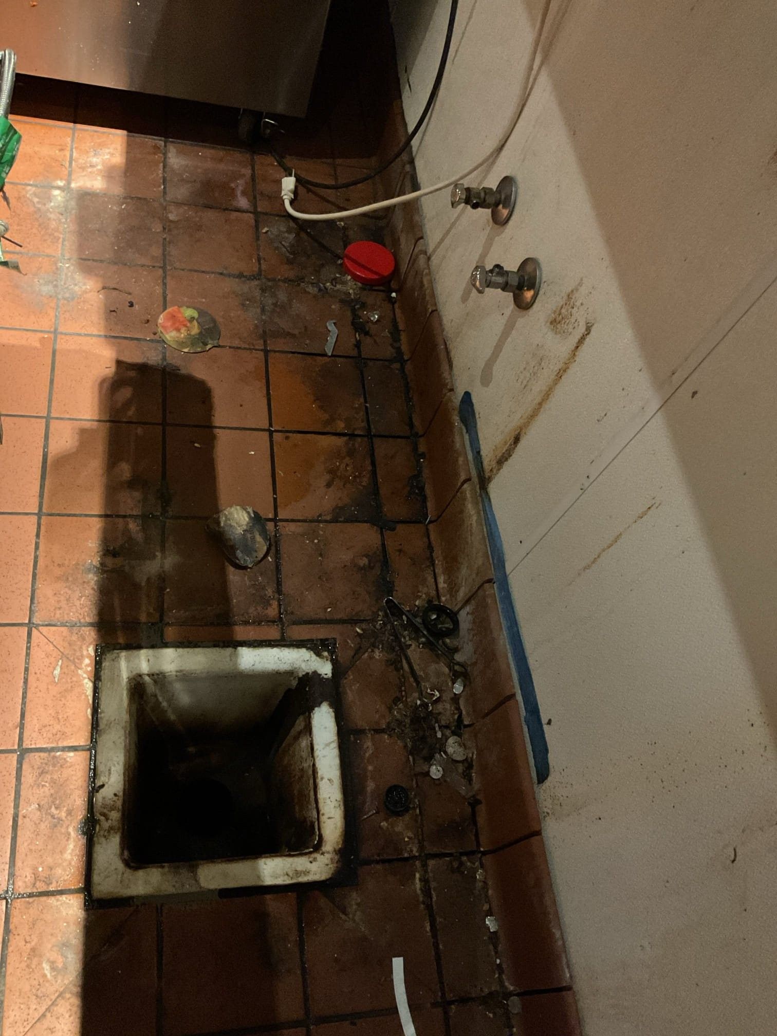 Dirty kitchen floor with a floor drain. Brown tiles, white walls, and pipes.