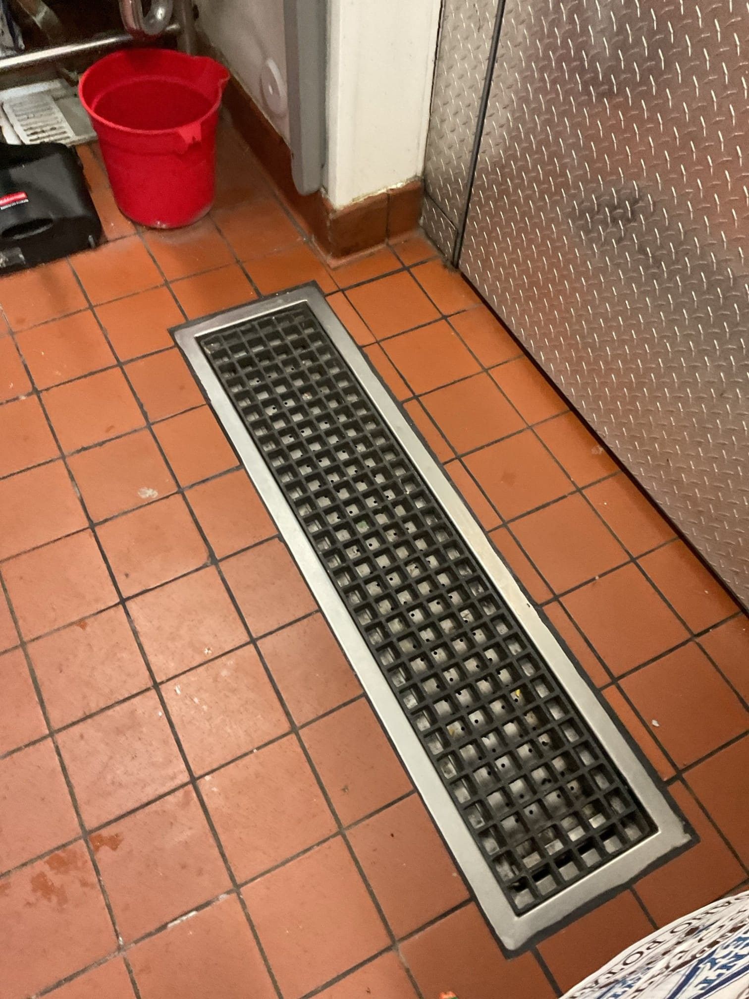 Floor drain with a metal grate on red tile in a commercial space. A red bucket and metal door are nearby.